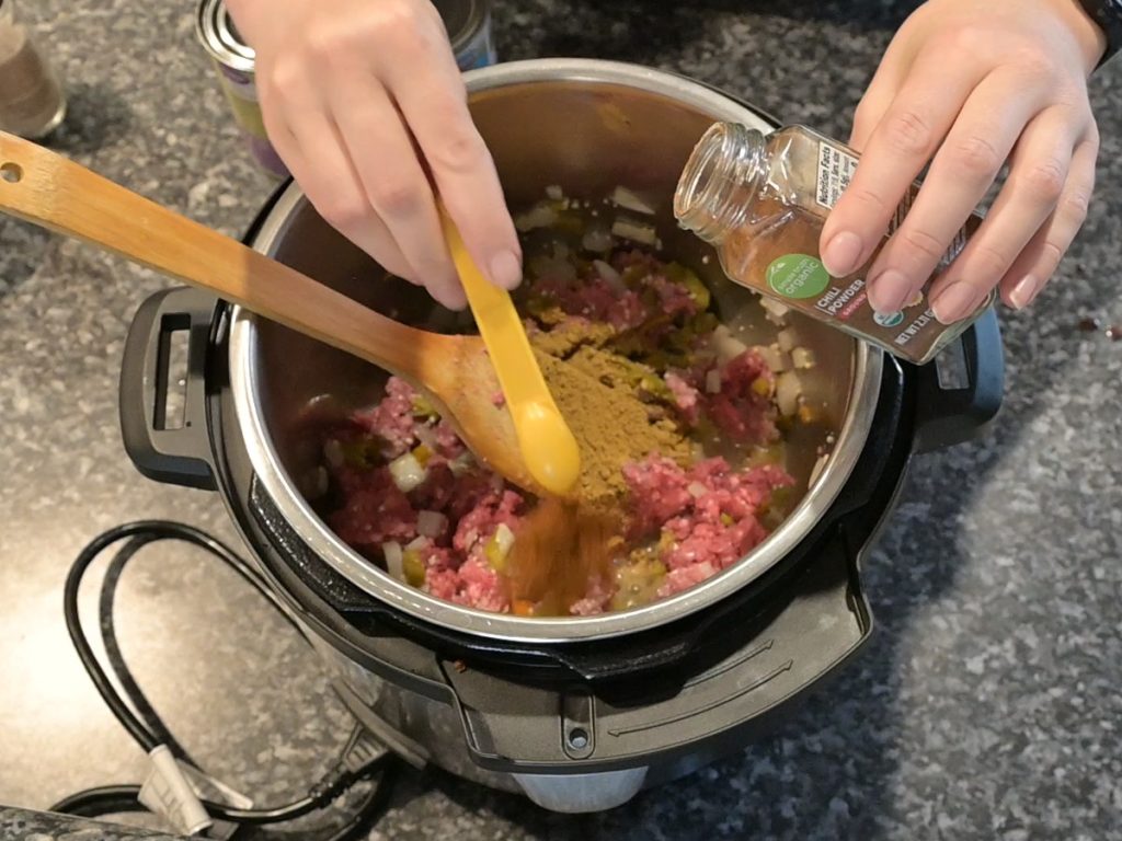 adding chili powder to ground beef, onions, and green chilis in the instant pot